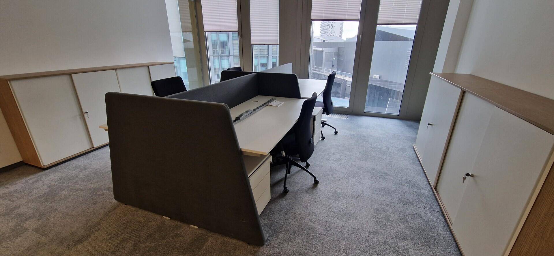 Heller Bürobereich mit mehreren Arbeitsplätzen, Teppichboden und raumhohen Fenstern.