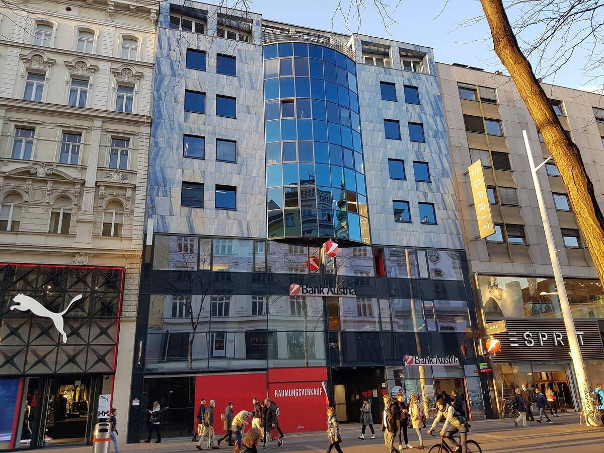 Moderne Geschäftsfassade mit Glas- und Steinelementen an einer belebten Einkaufsstraße, umgeben von älteren Gebäuden.