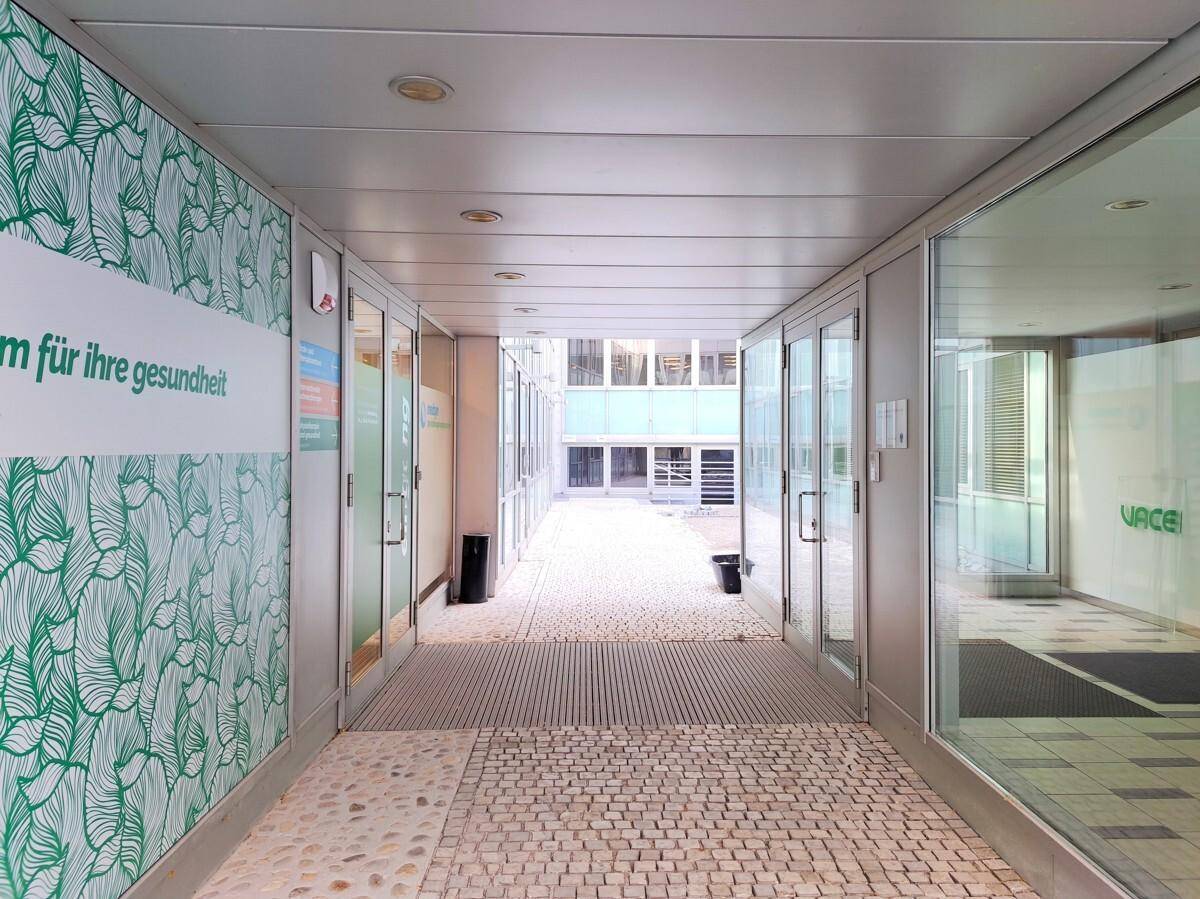 Moderner Flur mit Glaswänden, die zu Büroräumen führen, und einem gepflasterten Bodenbelag.