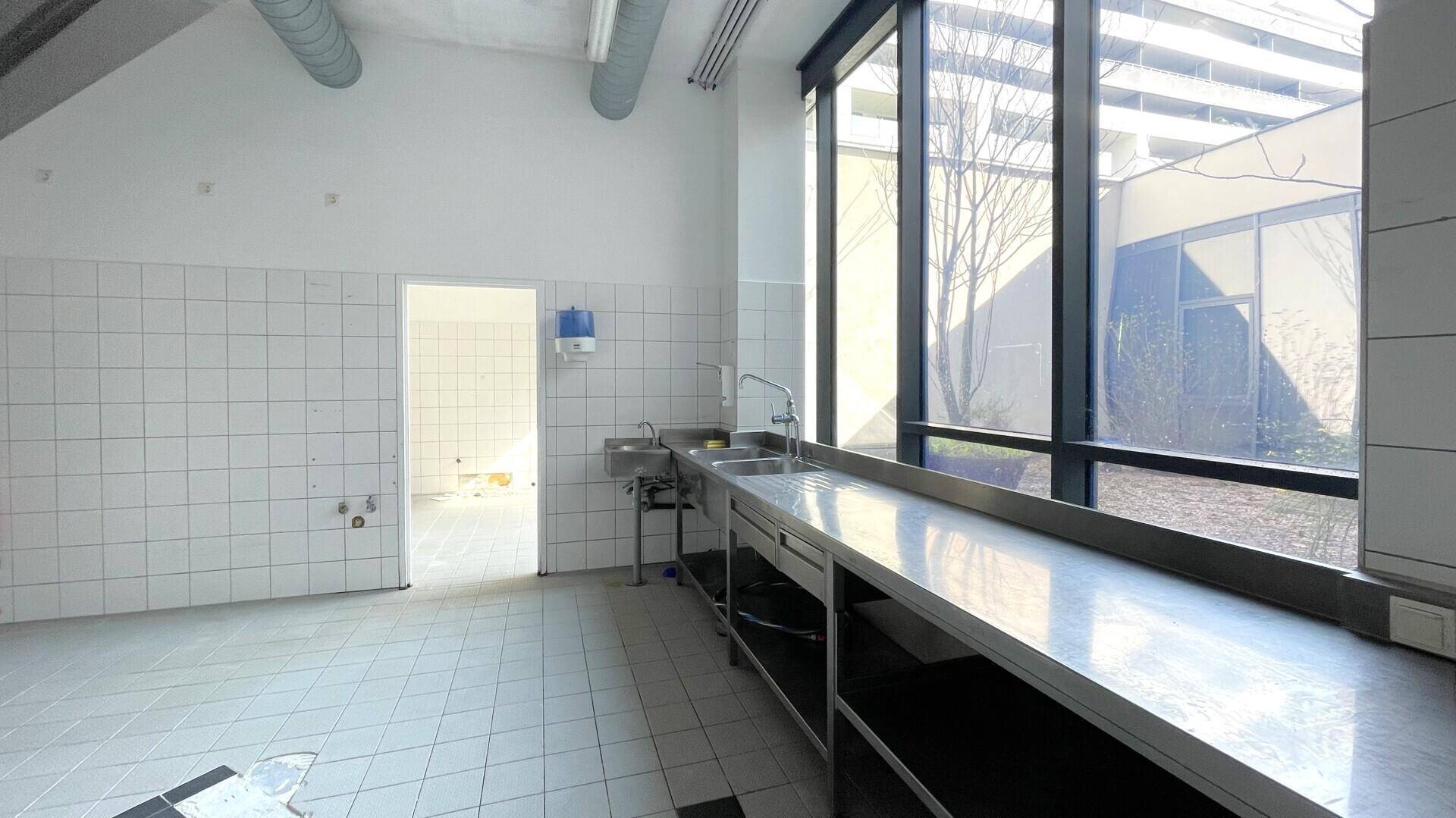 Heller Küchenbereich mit großen Fenstern, Edelstahlarbeitsflächen und gefliesten Wänden.