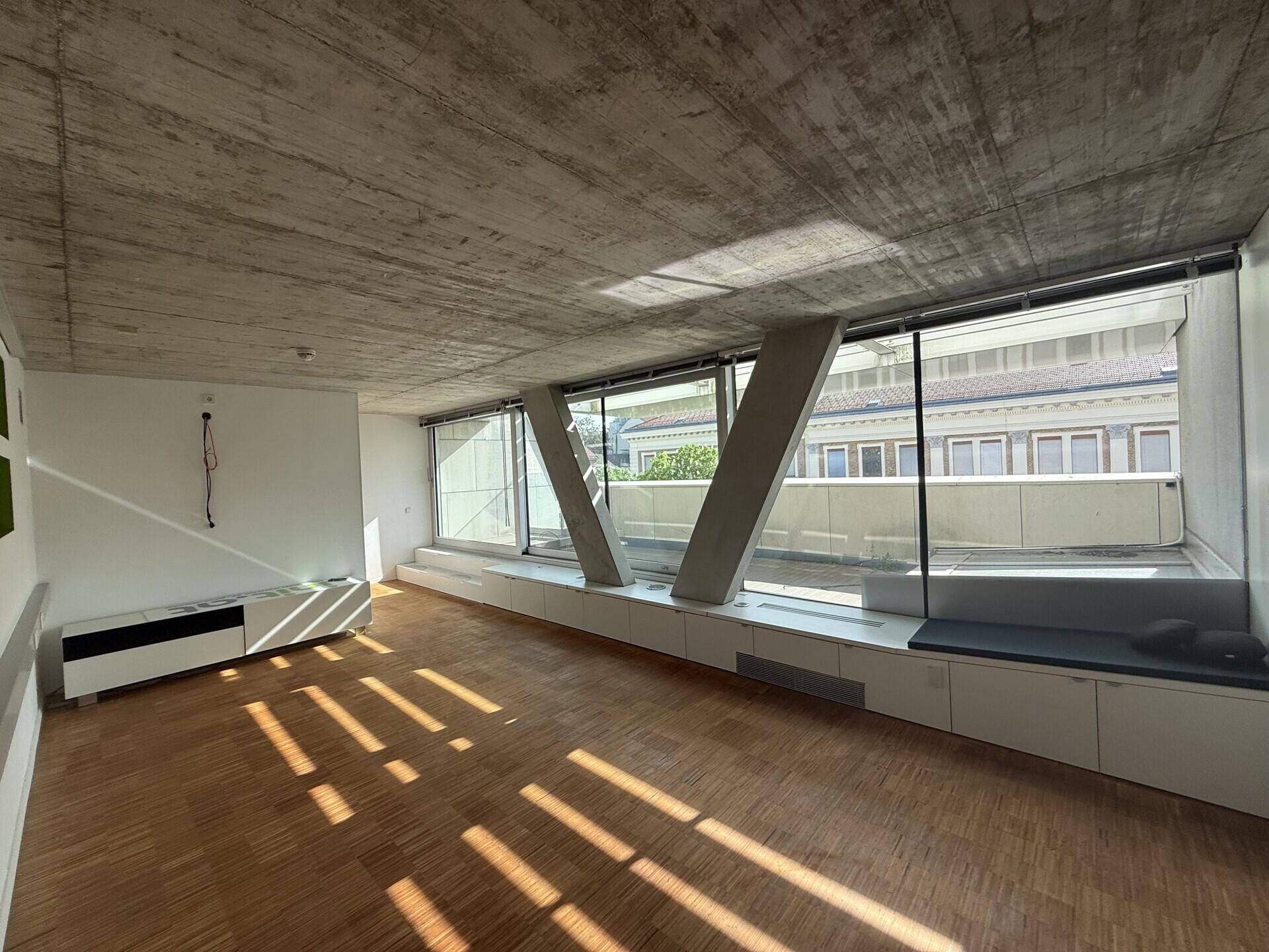 Heller Büroraum mit Holzboden, Sichtbetondecke und großen Fenstern, die viel Tageslicht hereinlassen.