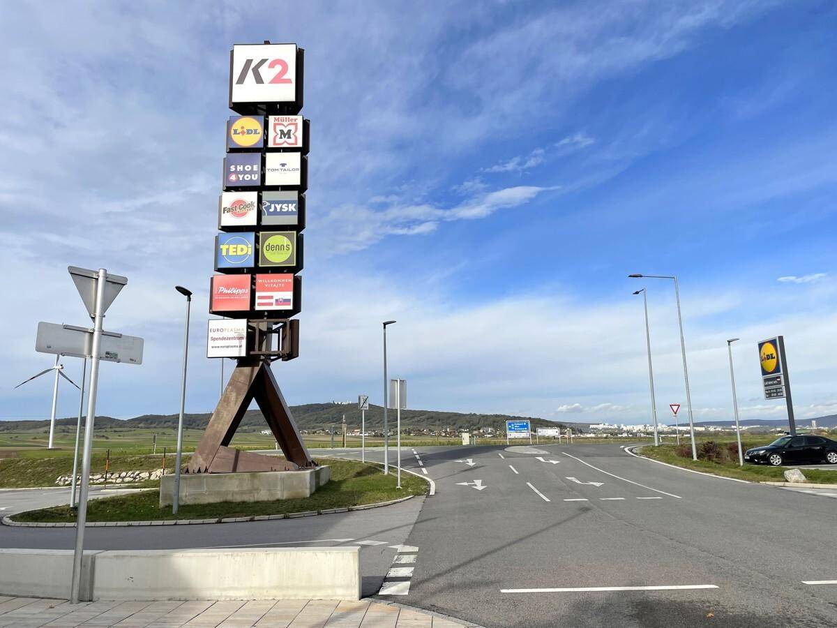 Hoher Werbepylon mit verschiedenen Geschäftslogos an einer Zufahrtsstraße zu einem Einkaufszentrum unter blauem Himmel.