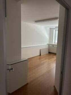 Blick durch eine Tür in einen Büroraum mit Holzboden, weißen Wänden und einem Fenster mit Jalousien.