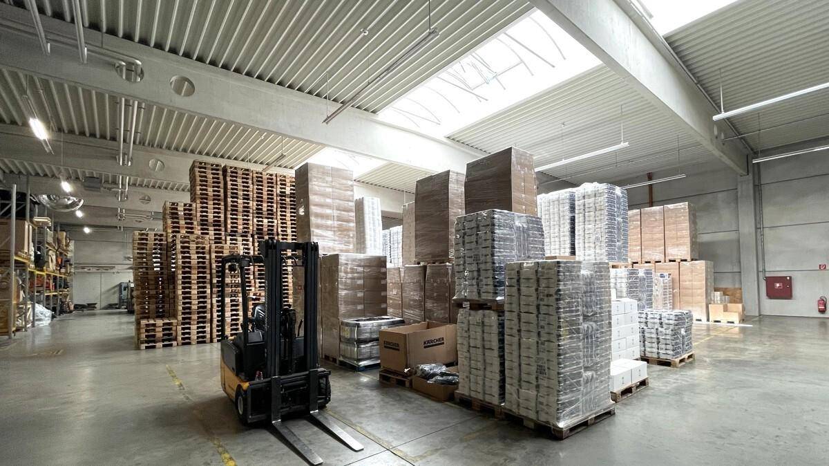 Großes Lager mit gestapelten Paletten, verpackten Waren und einem Gabelstapler unter Oberlichtern.