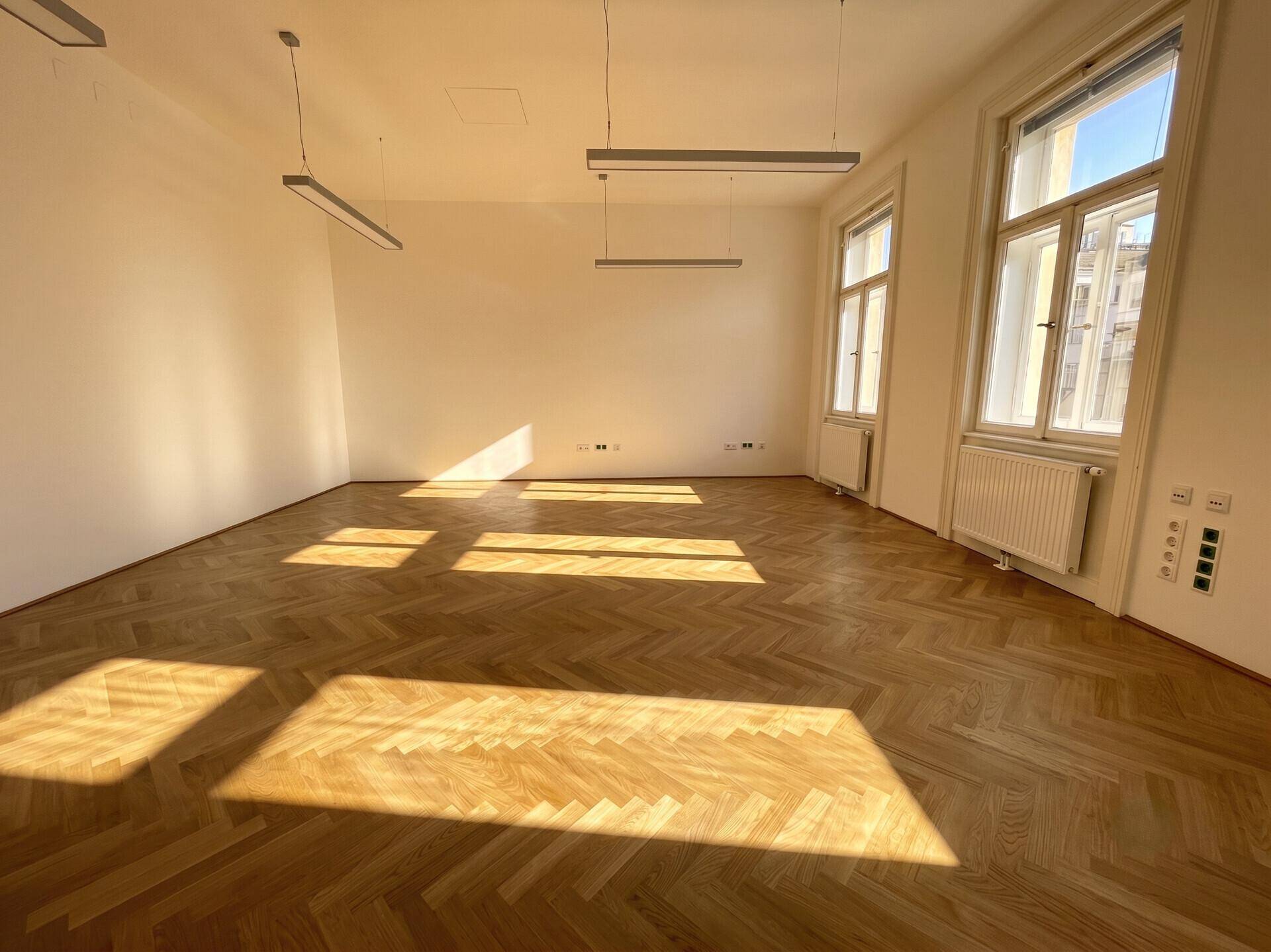 Geräumiger Büroraum mit hellem Fischgrätparkett und modernen Deckenleuchten, durchflutet von Sonnenlicht.