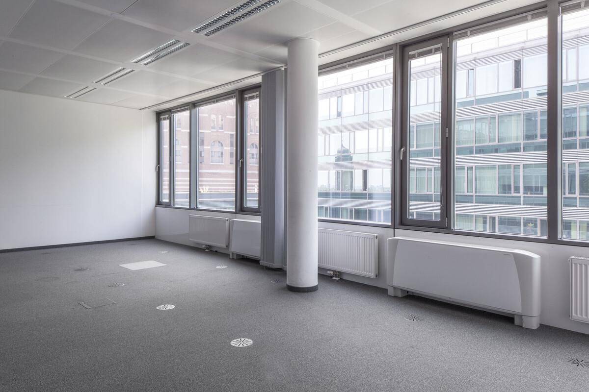 Geräumiger, leerer Büroraum mit großen Fenstern, die viel Tageslicht hereinlassen, und einem Teppichboden.