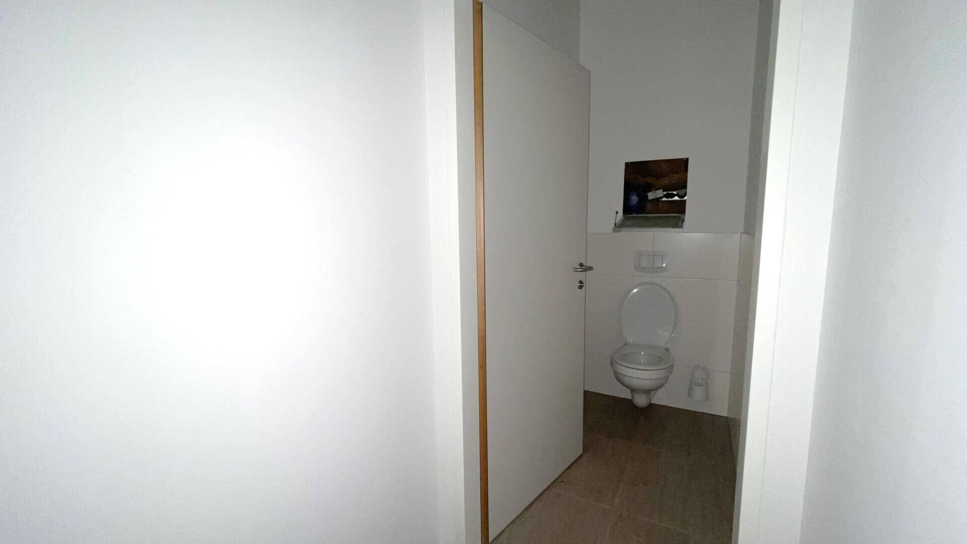 Einfache Toilette mit weiß gefliesten Wänden, WC und einem kleinen Fenster oder einer Öffnung über dem Spülkasten.