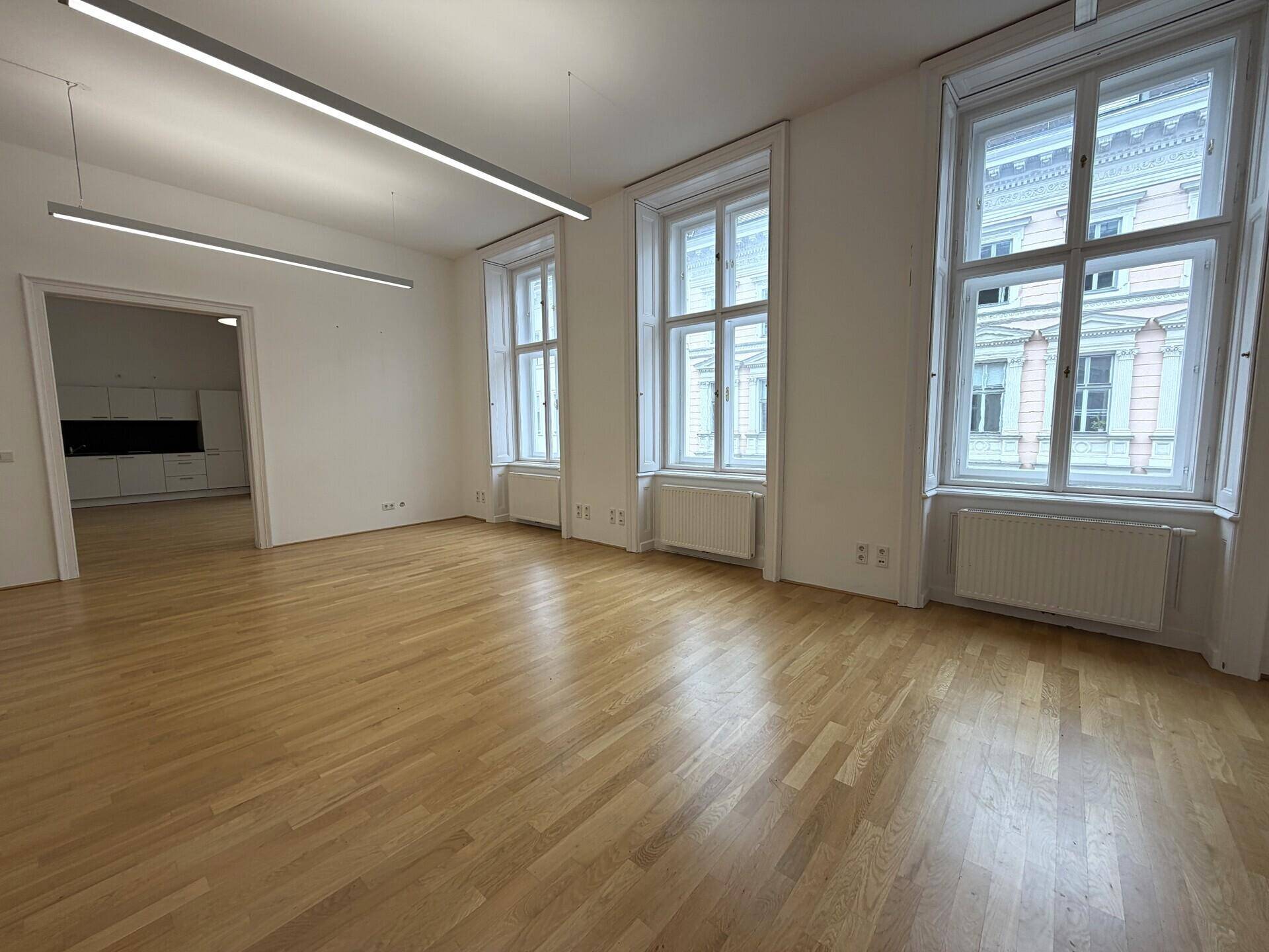 Geräumiges Büro mit hellem Holzparkett, großen Fenstern und moderner Deckenbeleuchtung.