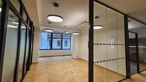 Helles Büro mit Fischgrätparkett, Glaswänden und modernen Deckenleuchten, das viel Tageslicht bietet.