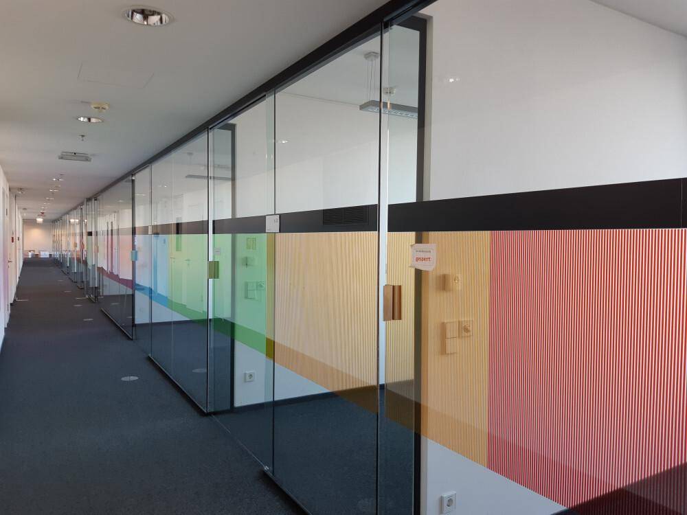 Langer Büroflur mit Glaswänden, teilweise farbiger Folierung und dunklem Teppichboden.