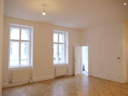 Helles Zimmer mit zwei großen Fenstern, Parkettboden und Heizkörpern unter den Fenstern.
