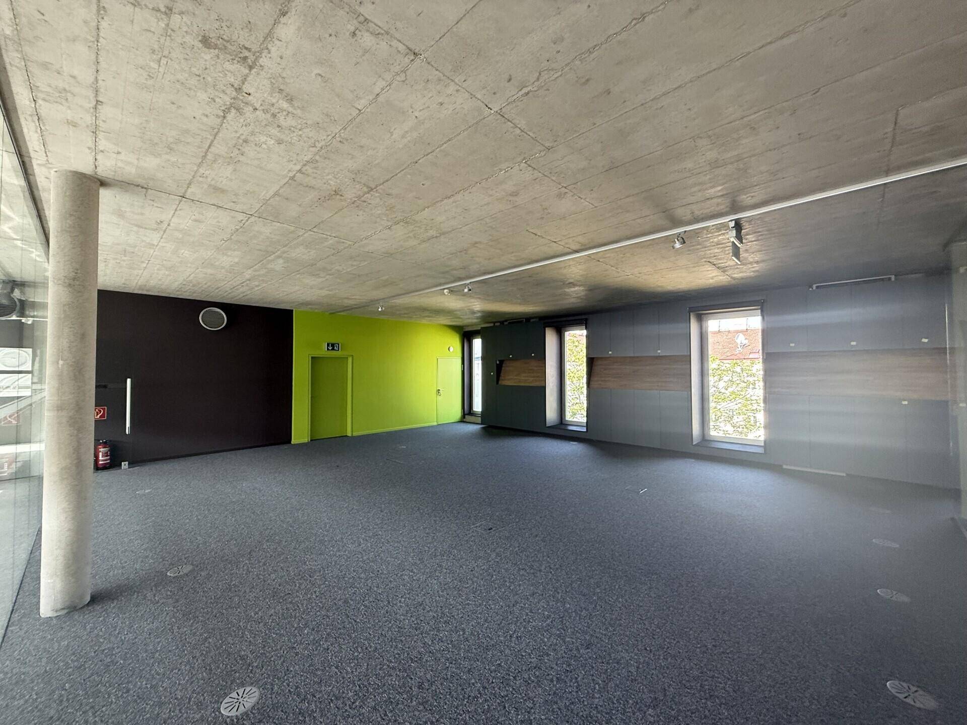 Offener Bürobereich mit Teppichboden, Sichtbetondecke und farbigen Akzentwänden für eine moderne Atmosphäre.