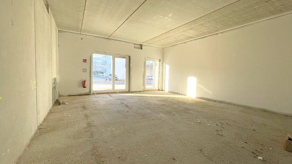 Großer, heller Gewerberaum im Rohbau mit Betonboden und großen Fenstern, die viel Licht hereinlassen.