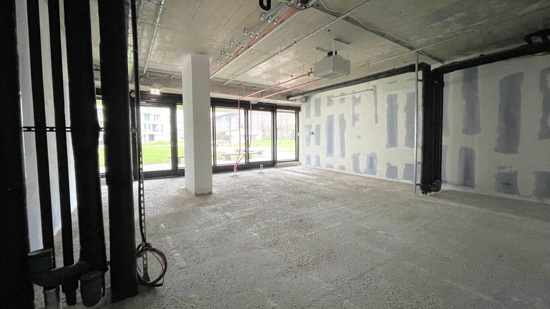 Großer, unfertiger Gewerberaum mit Betonboden, freiliegenden Deckeninstallationen und bodentiefen Fenstern zum Außenbereich.
