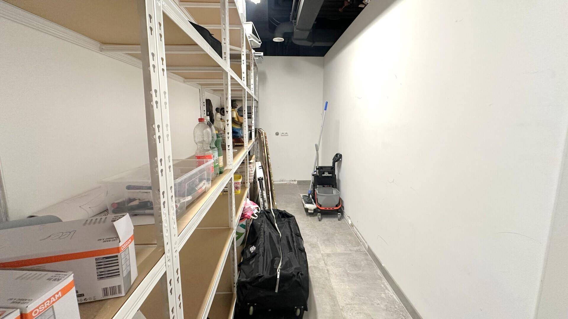 Schmaler Lagerraum mit Regalen an der Wand und Reinigungsgeräten am Ende des Ganges.