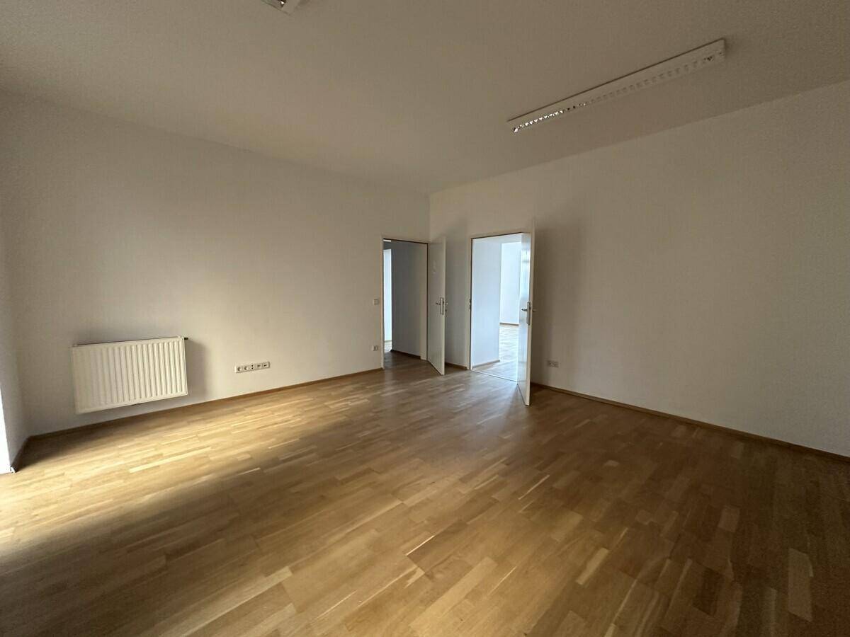 Geräumiger Büroraum mit hellem Parkettboden, weißen Wänden und mehreren Türen zu angrenzenden Bereichen.
