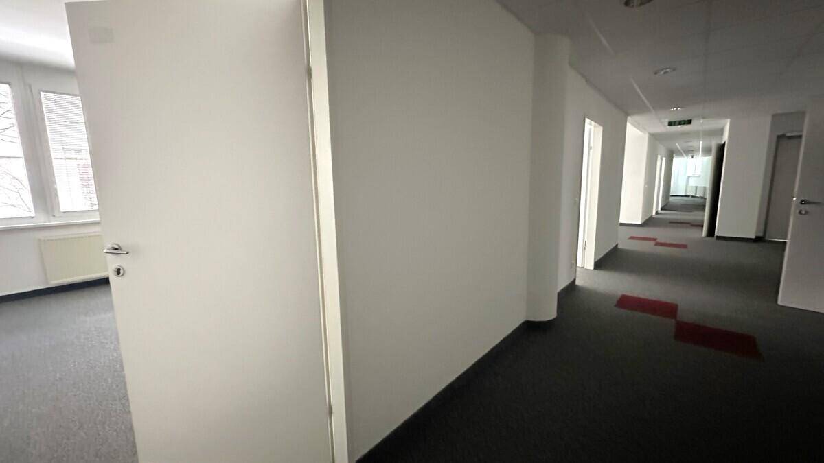 Langer Bürogang mit grauem Teppichboden, weißen Wänden und mehreren Türen zu den einzelnen Büros.