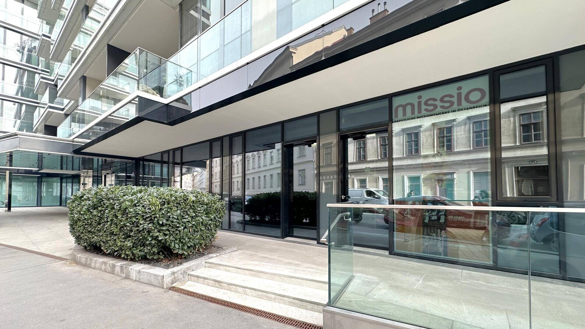Ansicht der modernen Gebäudefassade mit großen Glasflächen und einem begrünten Eingangsbereich an der Straße.