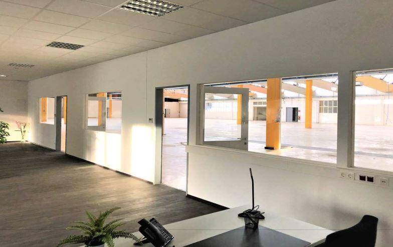Büroraum mit dunklem Boden und weißen Wänden, der durch große Fenster einen Blick in eine Lagerhalle bietet.