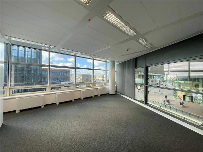 Geräumiges, leeres Büro mit Teppichboden und großen Fenstern, die viel Tageslicht hereinlassen und einen Stadtblick bieten.