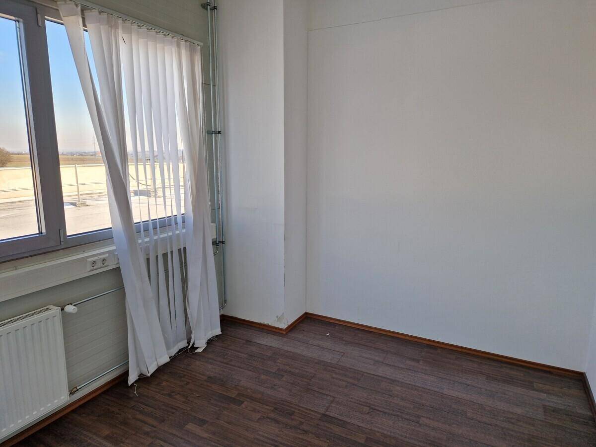 Leerer Raum mit dunklem Laminatboden, einem Fenster mit vertikalen Jalousien und einem Heizkörper.