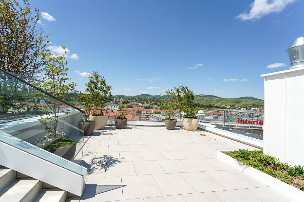 Geräumige Dachterrasse mit moderner Glasbrüstung, Pflanzen in Töpfen und weitem Blick auf die Stadt und Hügel.
