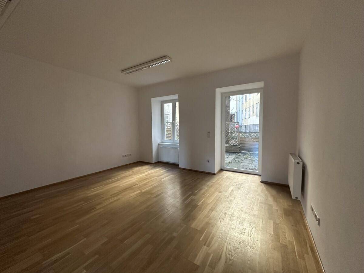 Heller Büroraum mit Parkettboden und Zugang zu einem kleinen Außenbereich durch eine Glastür.
