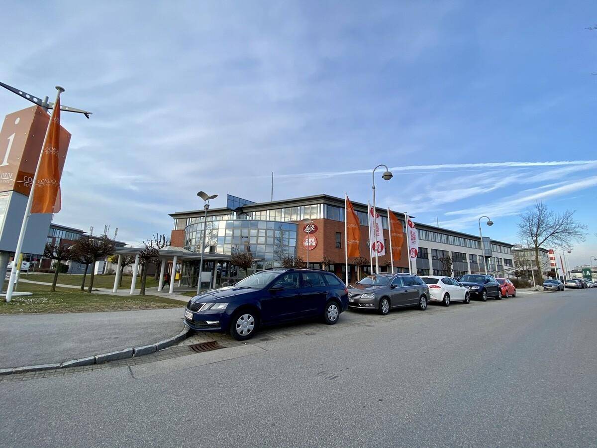 Blick auf ein großes Geschäftsgebäude mit moderner Glasfassade, umgeben von Parkplätzen und Fahnenmasten.