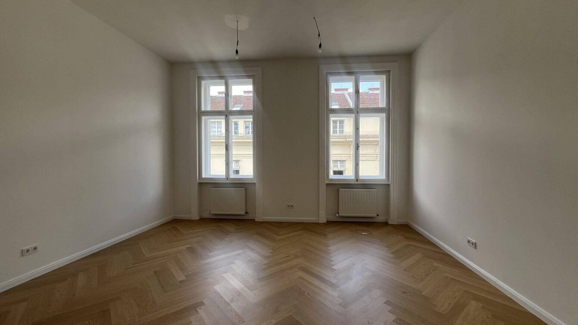 Heller Raum mit zwei hohen Fenstern, die viel Tageslicht hereinlassen, und elegantem Fischgrätparkettboden.