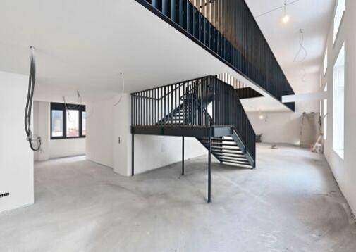 Geräumiger, unmöblierter Gewerberaum mit Betonboden, hohen Decken und einer modernen Metalltreppe.