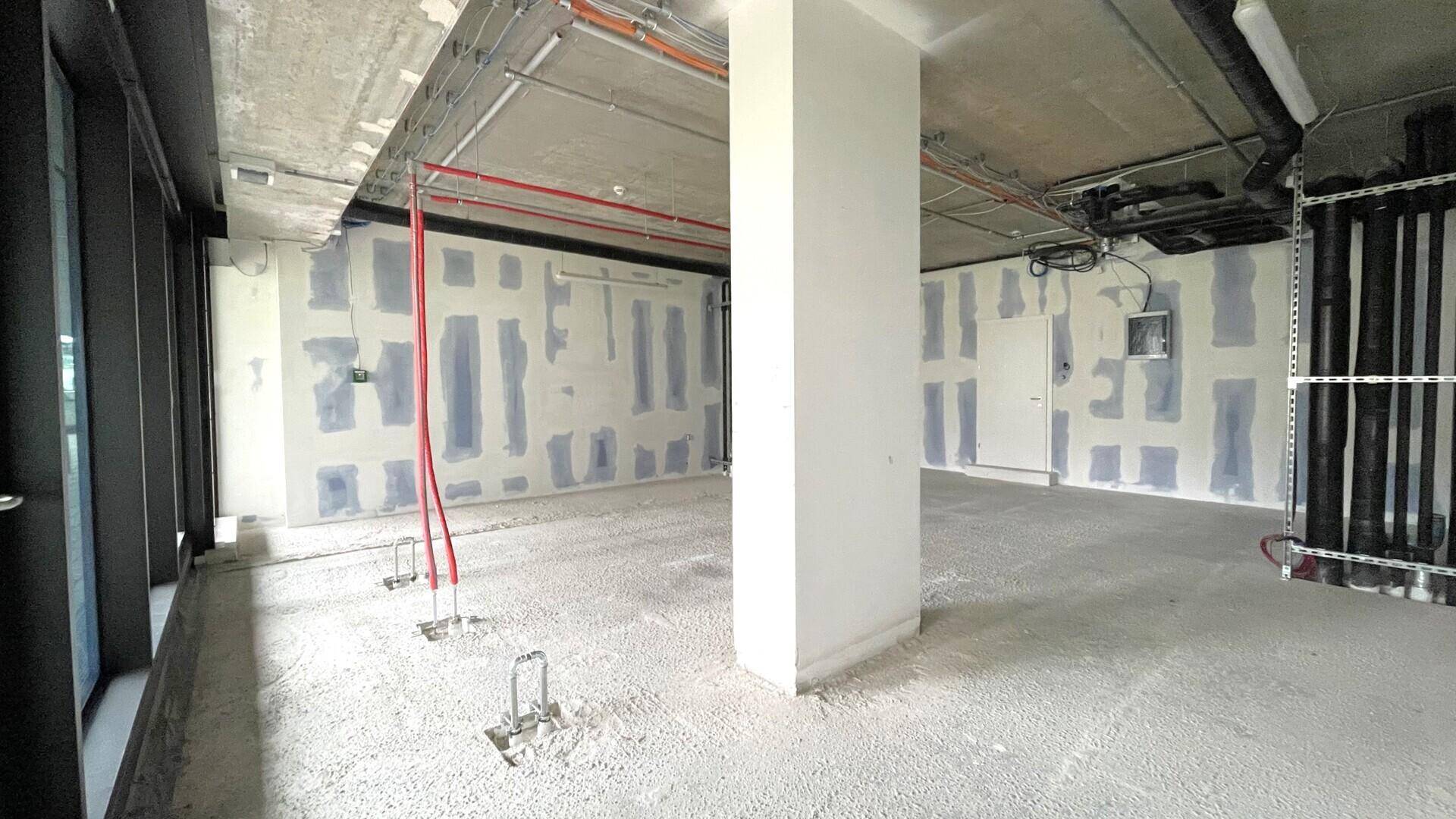 Unfertiger Gewerberaum mit freiliegenden Leitungen, Betonboden und großen Fenstern, bereit für den Innenausbau.