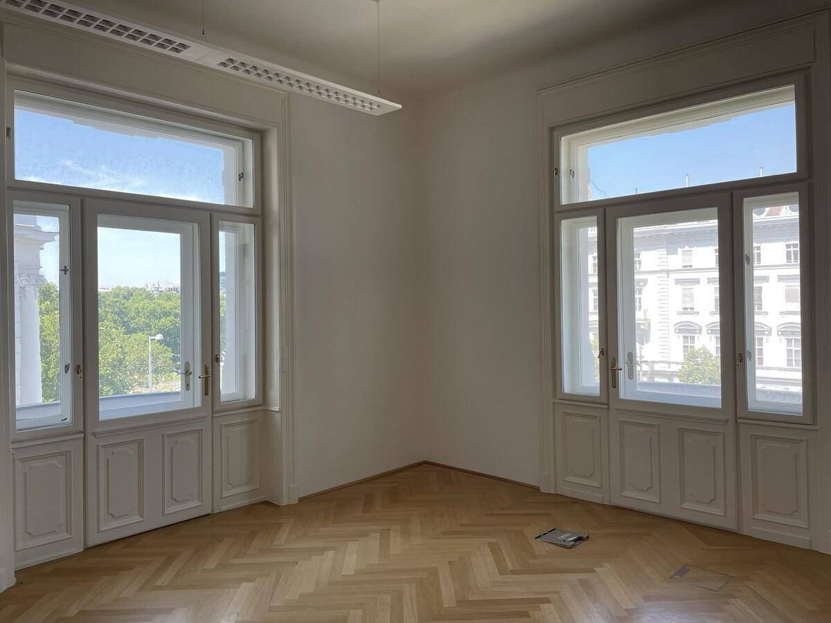 Großer, heller Raum mit Fischgrätparkett und hohen Fenstertüren, die viel Licht hereinlassen.