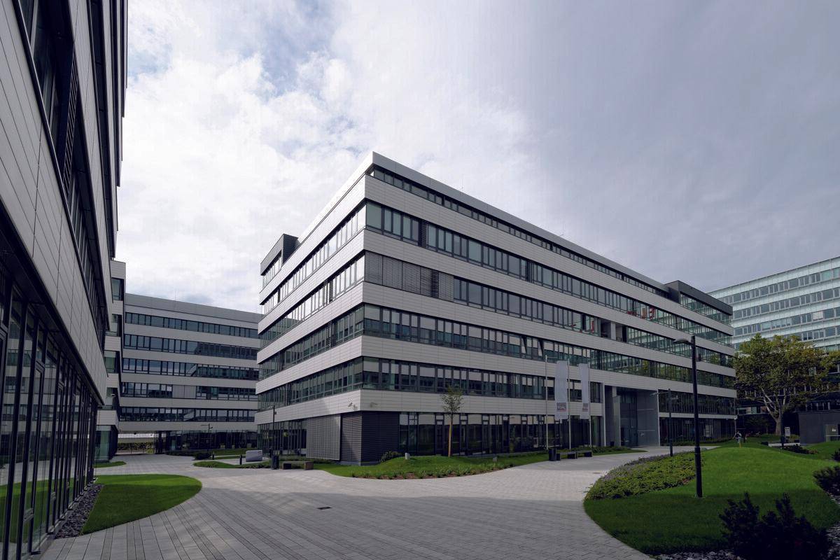 Mehrere moderne Bürogebäude mit großen Fensterfronten und gepflegten Außenanlagen unter bewölktem Himmel.