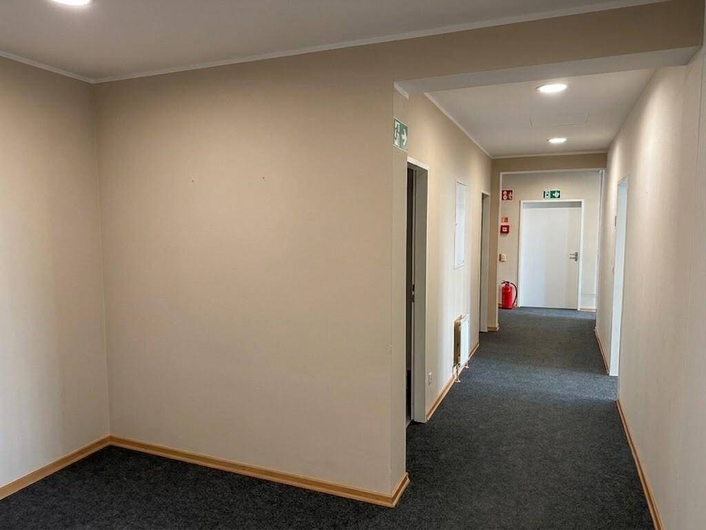 Flur mit Teppichboden und hellen Wänden, der zu weiteren Räumen und einem Notausgang führt.