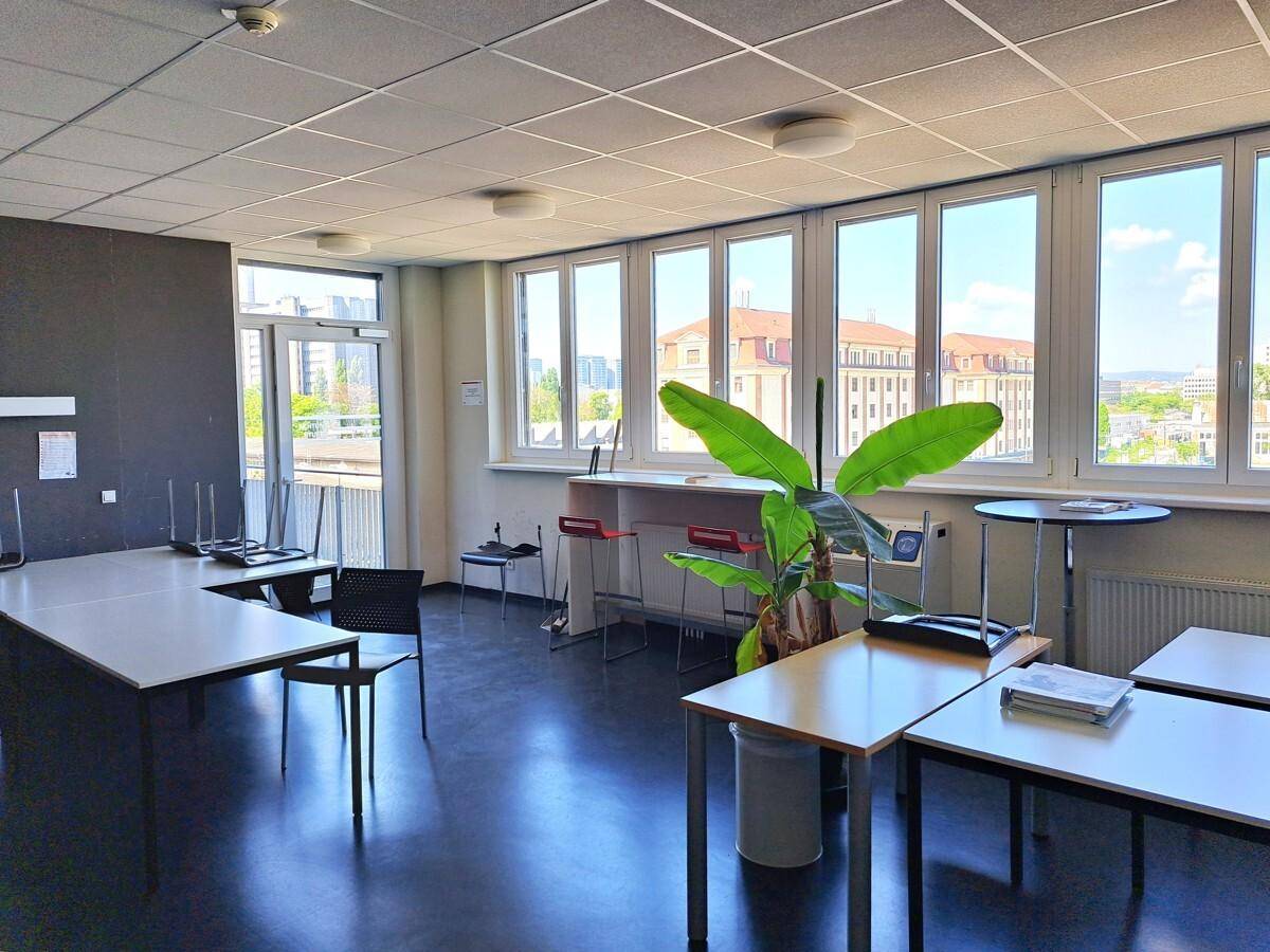 Geräumiger Gemeinschaftsraum mit Tischen, Stühlen und großen Fenstern, die viel Tageslicht hereinlassen.