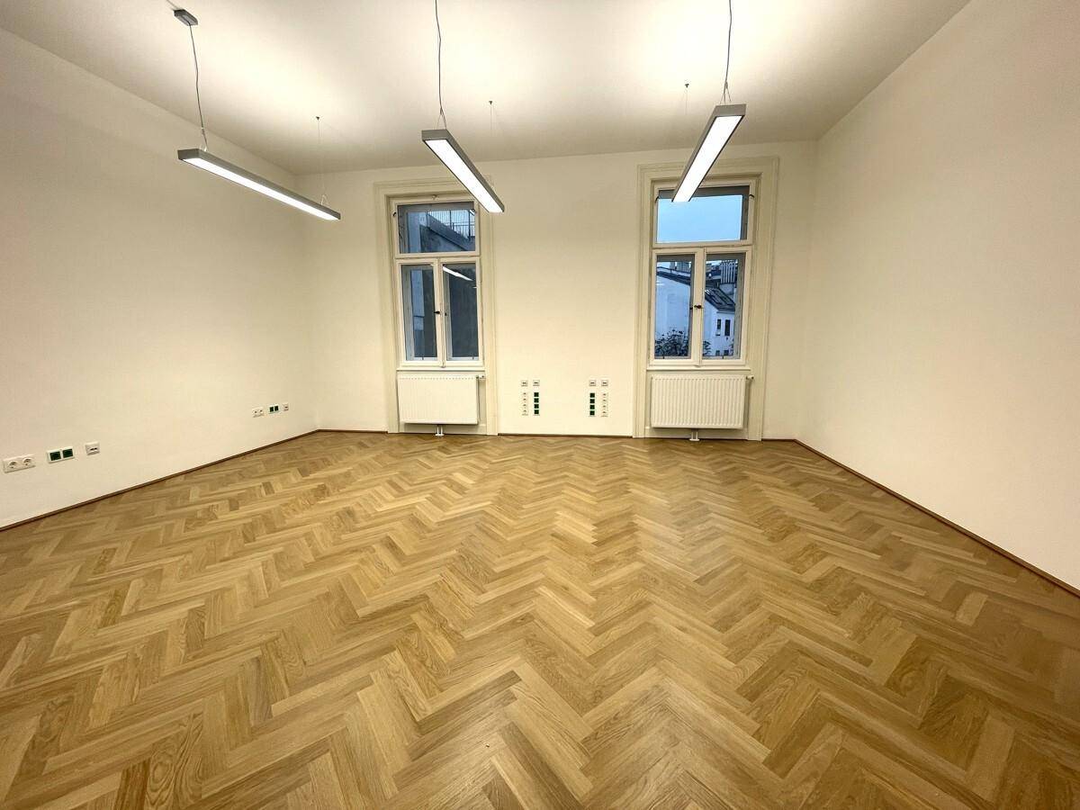 Geräumiger Büroraum mit Fischgrätparkett und zwei hohen Fenstern, die den Raum mit Tageslicht füllen.