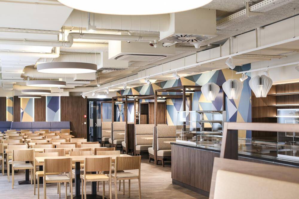 Geräumige Cafeteria mit vielen Sitzgelegenheiten, einer Essensausgabe und moderner Deckenbeleuchtung.