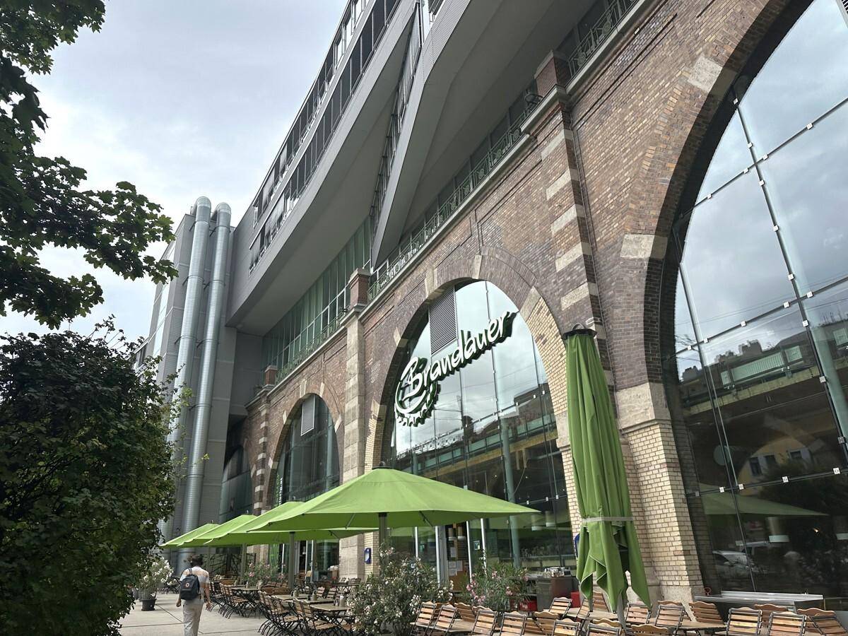 Außenansicht eines modernen Gebäudes mit Ziegelbögen, großer Glasfassade und einer Terrasse mit grünen Sonnenschirmen.