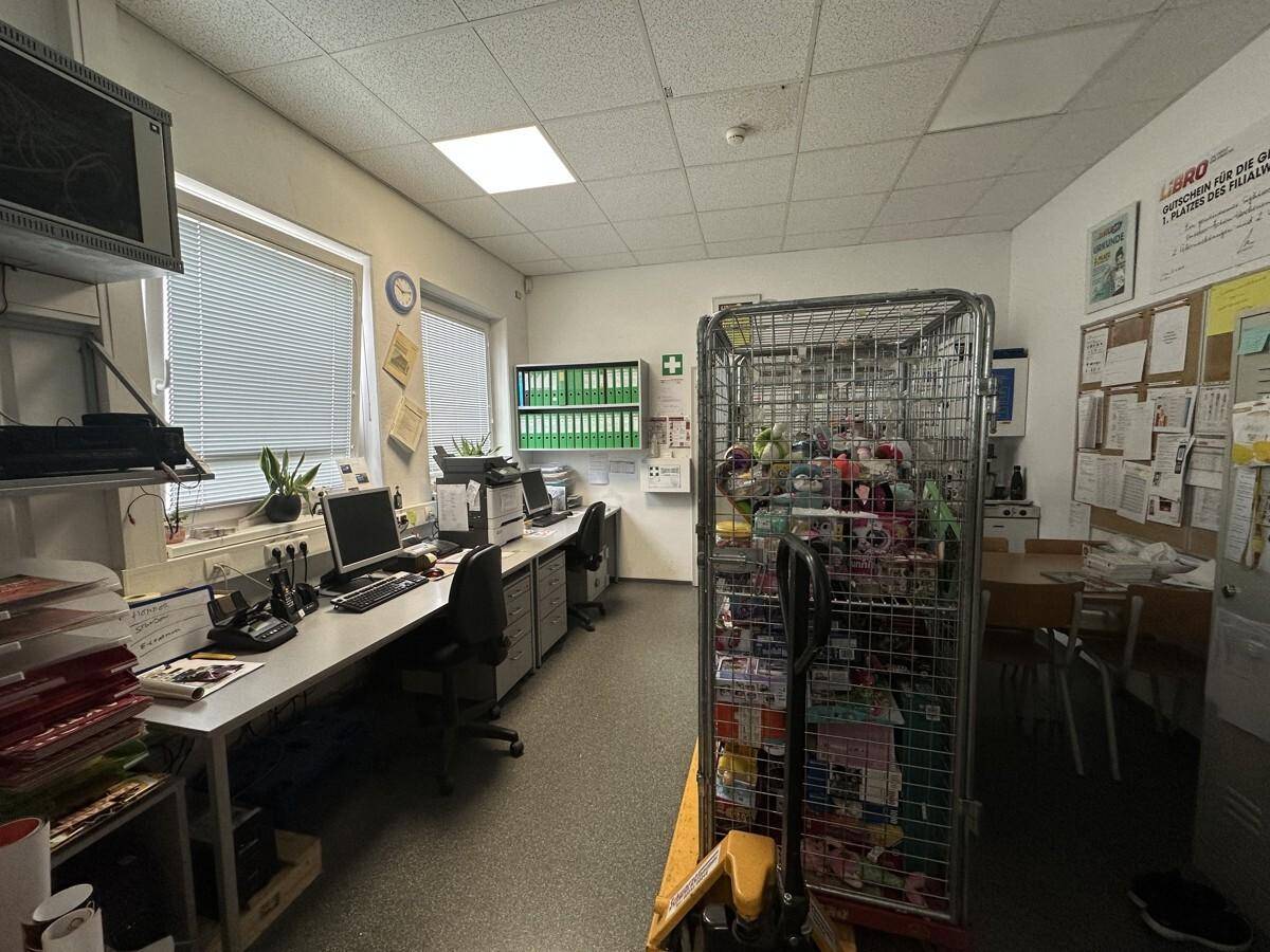 Büroraum mit mehreren Arbeitsplätzen, Computern und einem Rollcontainer, beleuchtet durch Deckenleuchten und Fenster.