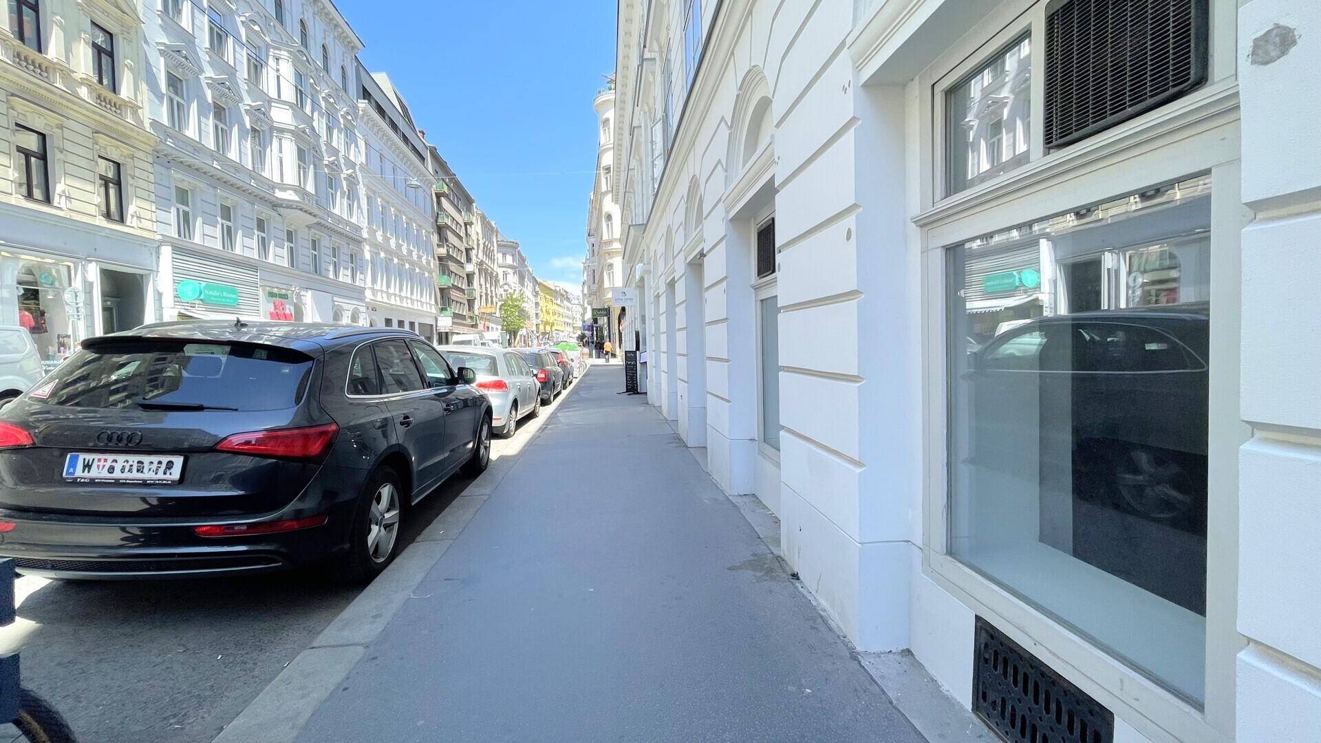 Blick entlang einer belebten Straße mit klassischen Gebäuden, geparkten Autos und einem breiten Bürgersteig.