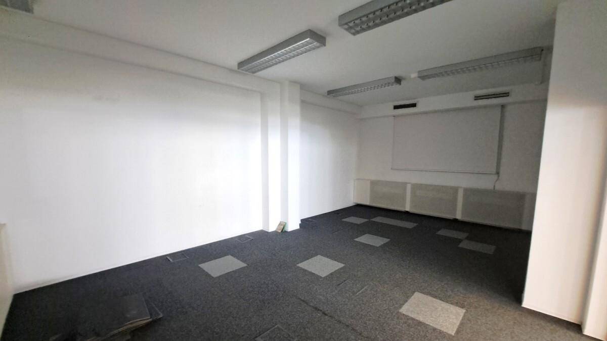 Büroraum mit Teppichfliesen, einer Projektionsfläche an der Wand und Deckenleuchten.