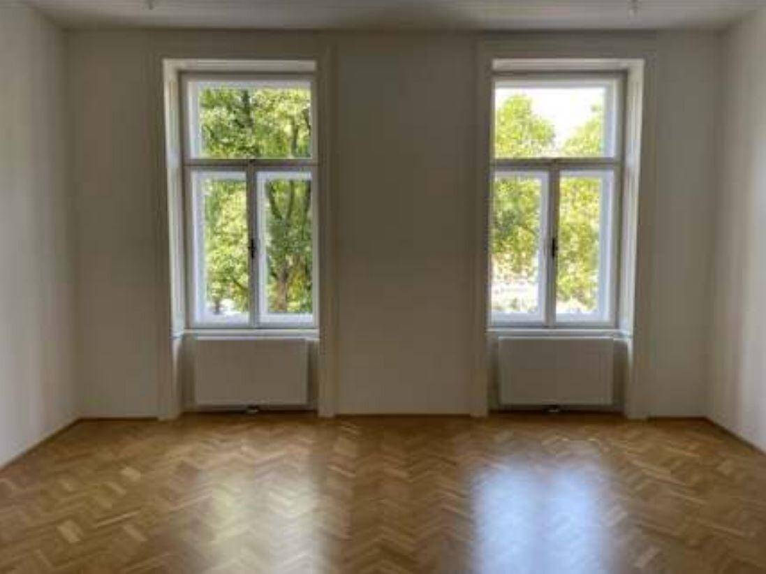Heller Raum mit Fischgrätparkett und zwei großen Fenstern mit Blick auf grüne Bäume.