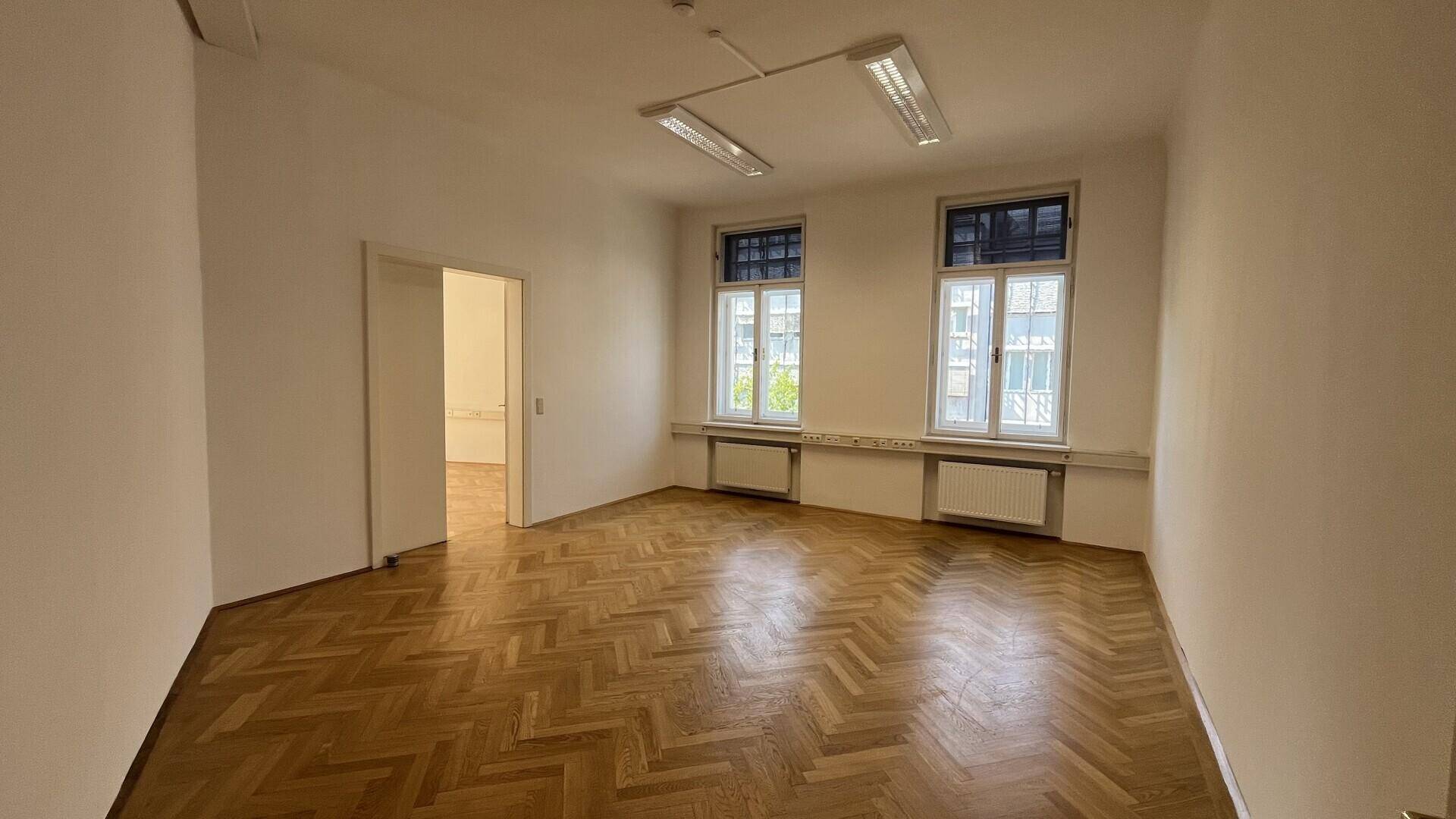 Helles Büro mit Fischgrätparkett, zwei Fenstern und Zugang zu einem weiteren Raum.