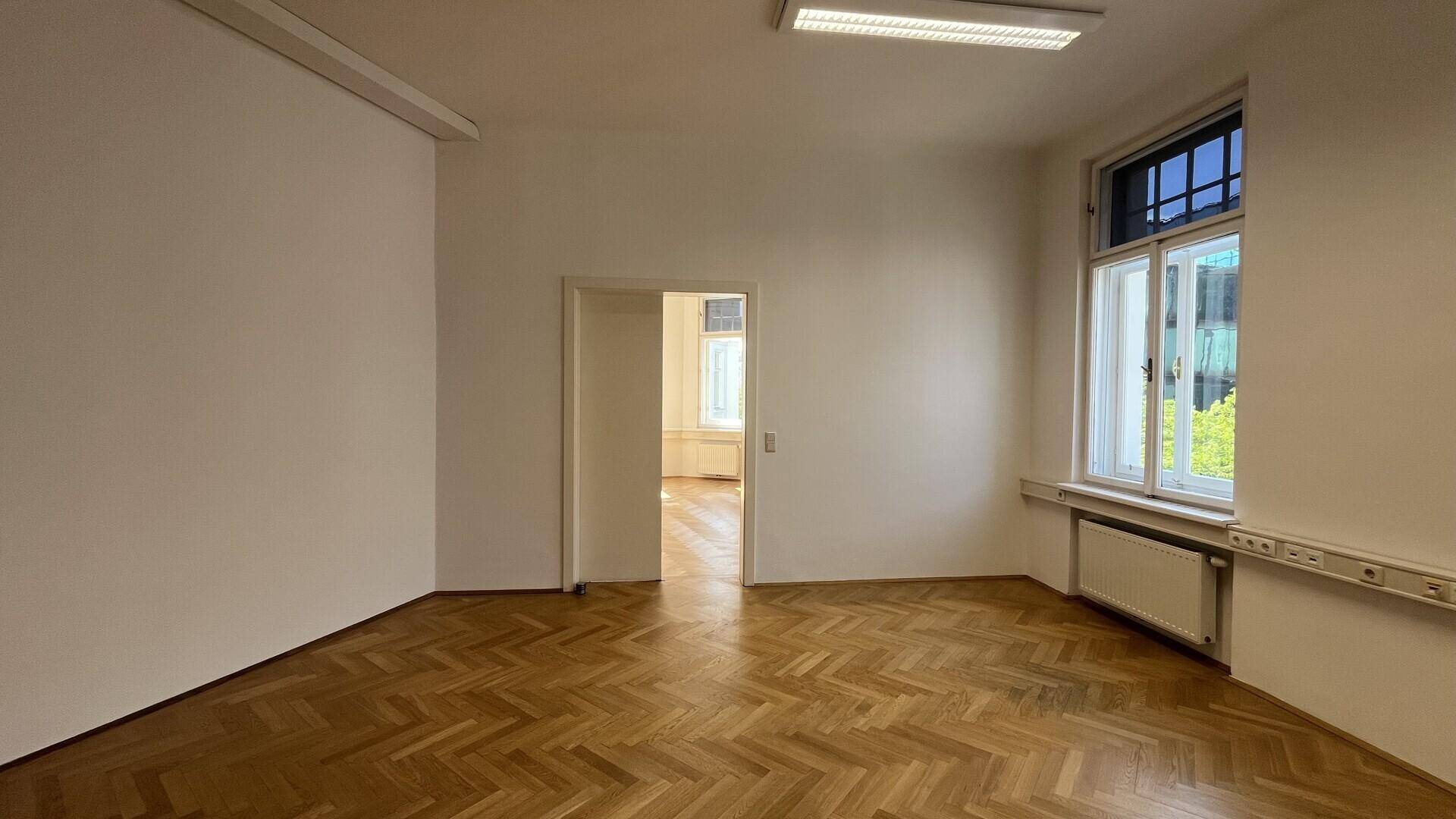 Leeres Büro mit Fischgrätparkett, einem großen Fenster und einer Verbindungstür zu einem anderen Raum.