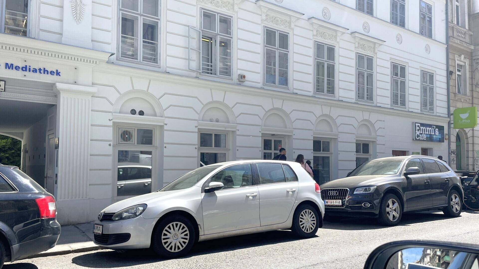 Nahaufnahme der weißen Fassade eines klassischen Gebäudes mit geparkten Autos und einem Café in der Nähe.