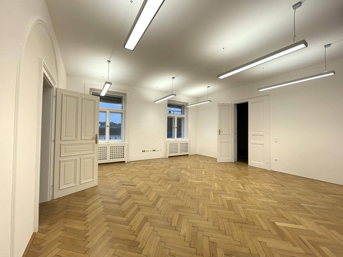 Großer Büroraum mit Fischgrätparkett, hohen Fenstern und klassischen Doppeltüren, die den Raum öffnen.