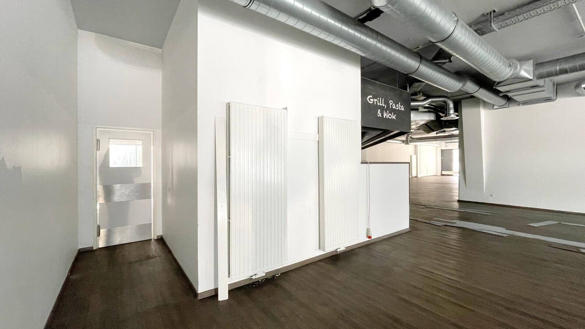 Leerer Gewerberaum mit Heizkörpern, einer Tür und sichtbaren Deckeninstallationen im Hintergrund.