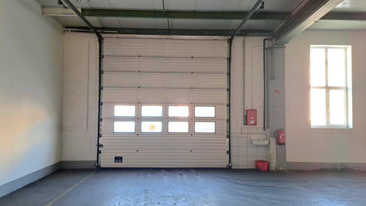 Innenansicht einer Lagerhalle mit großem Rolltor, Fenstern und Betonboden, gut beleuchtet.