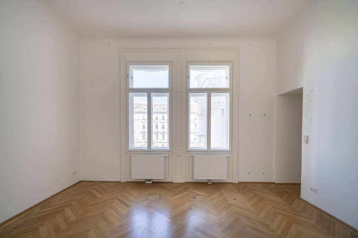 Heller, leerer Raum mit Fischgrätparkett und zwei großen Fenstern, die viel Tageslicht hereinlassen.