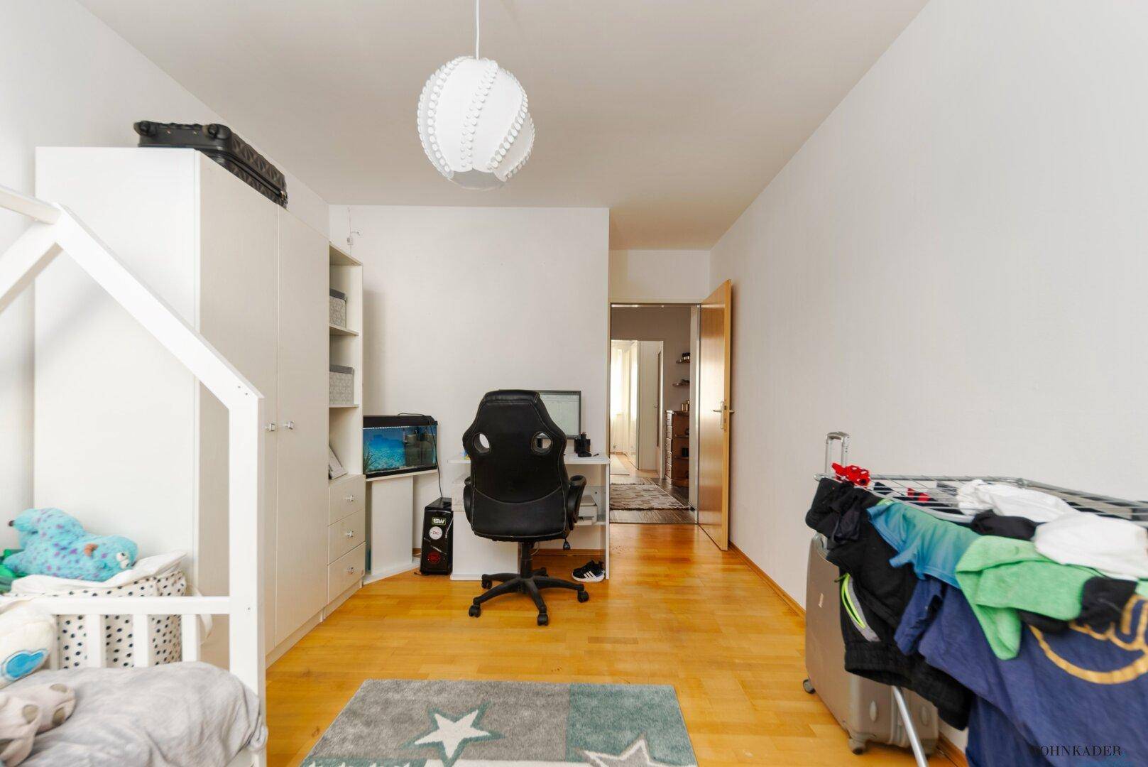 Geräumiges Kinderzimmer mit hellem Holzboden, Schreibtisch und einem kleinen Bett.